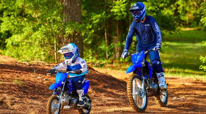 Image of father standing while riding his dirt bike looking at his son before him riding his Yamaha TTR50 dirt bike with trees at their back.